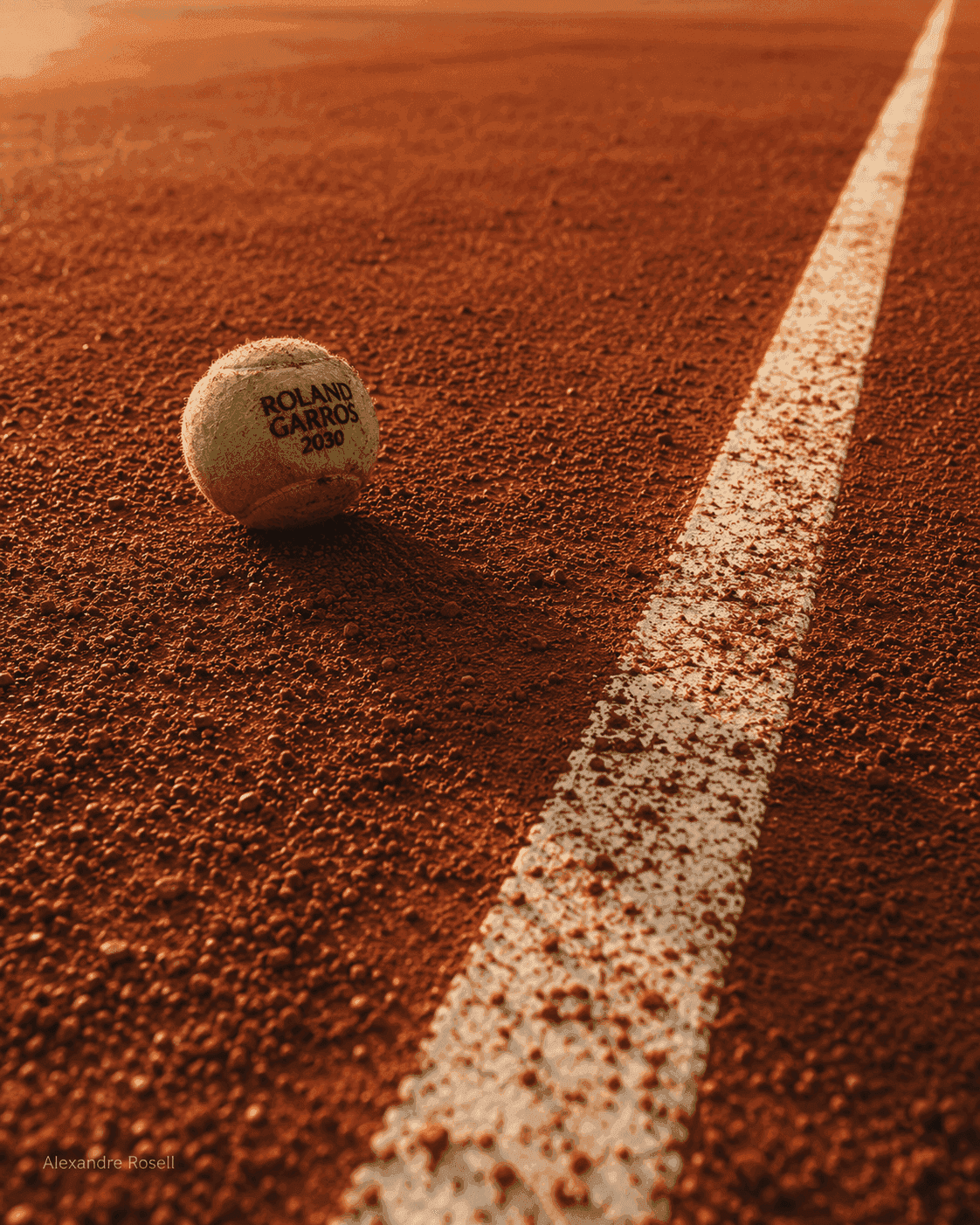A single ball on the white line of a red clay court in low evening light, marked Roland Garros.
