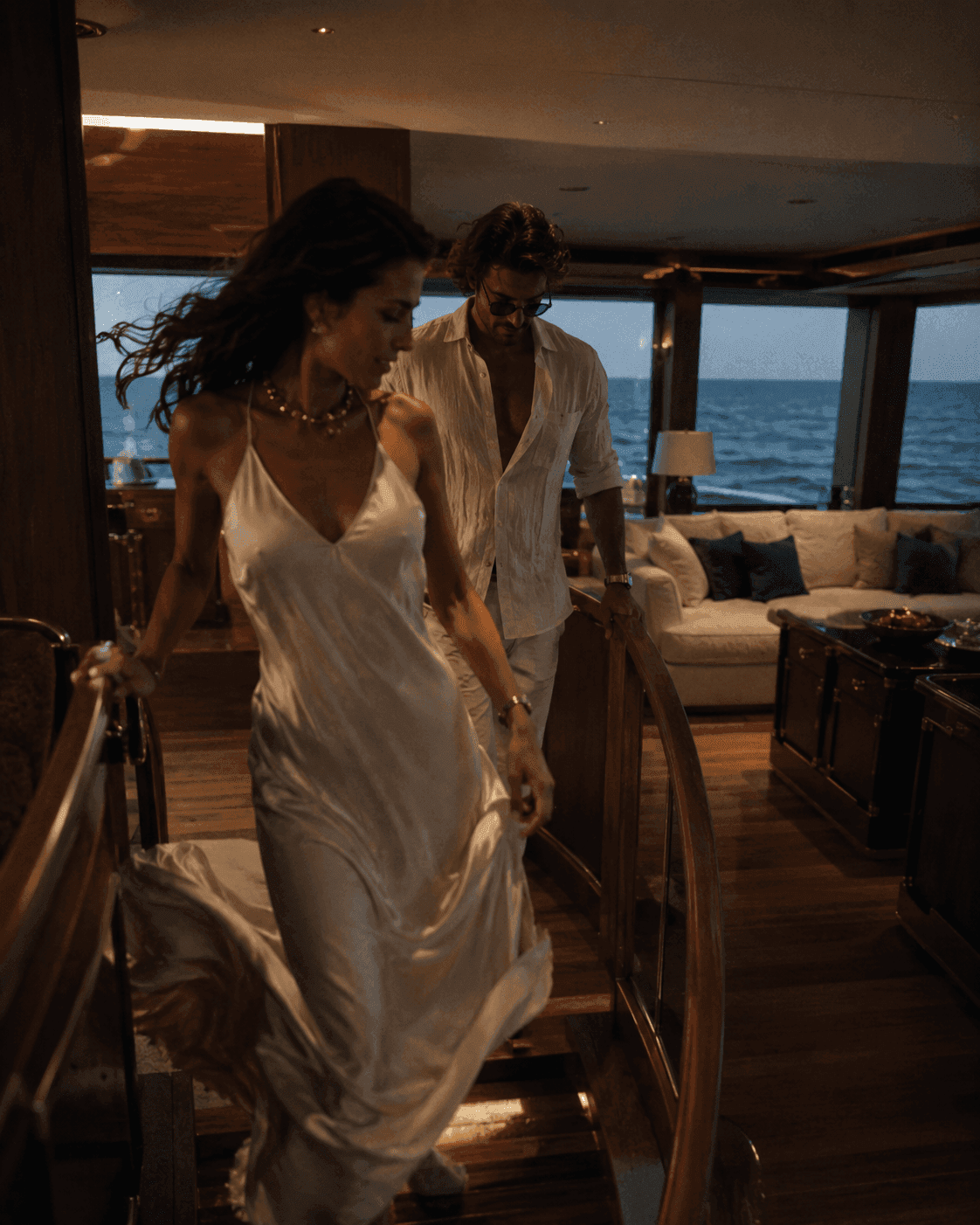 A couple in evening white moving through the warm-lit saloon of a yacht, the open sea beyond.